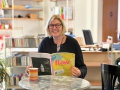 Eine Person sitzt im Lesecafé in der Mediathek und liest eine Zeitschrift. Sie lächelt in die Kamera.