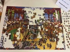 Auf einem Tisch liegt ein noch nicht fertiges Puzzle. Daneben steht ein SChild. Dort steht "Puzzeln Sie mit".