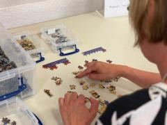 Eine Person sitzt am Puzzletisch und puzzelt. Das Bild ist von hinten über ihre Schulter fotografiert.