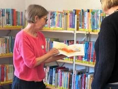 Eine Mitarbeiterin zeigt einer Kundin vor einem Regal in der Mediathek ein Buch.