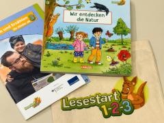 Auf einem Tisch liegt ein Buch, eine Stofftasche und ein Prospekt. Auf dem Bilderbuch steht "Wir entdecken die Natur".