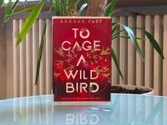 Das Buch To Cage a Wild Bird steht auf einem Glastisch. Im Hintergrund ist eine Pflanze.