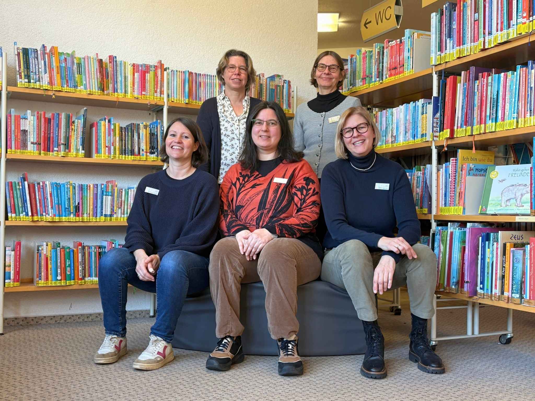 Das Mediatheksteam hat sich in der Mediathek f&uuml;r ein Foto aufgestellt. Vorne sitzen drei Frauen auf einem Hocker. Dahinter stehen zwei Frauen. Sie alle l&auml;cheln in die Kamera.