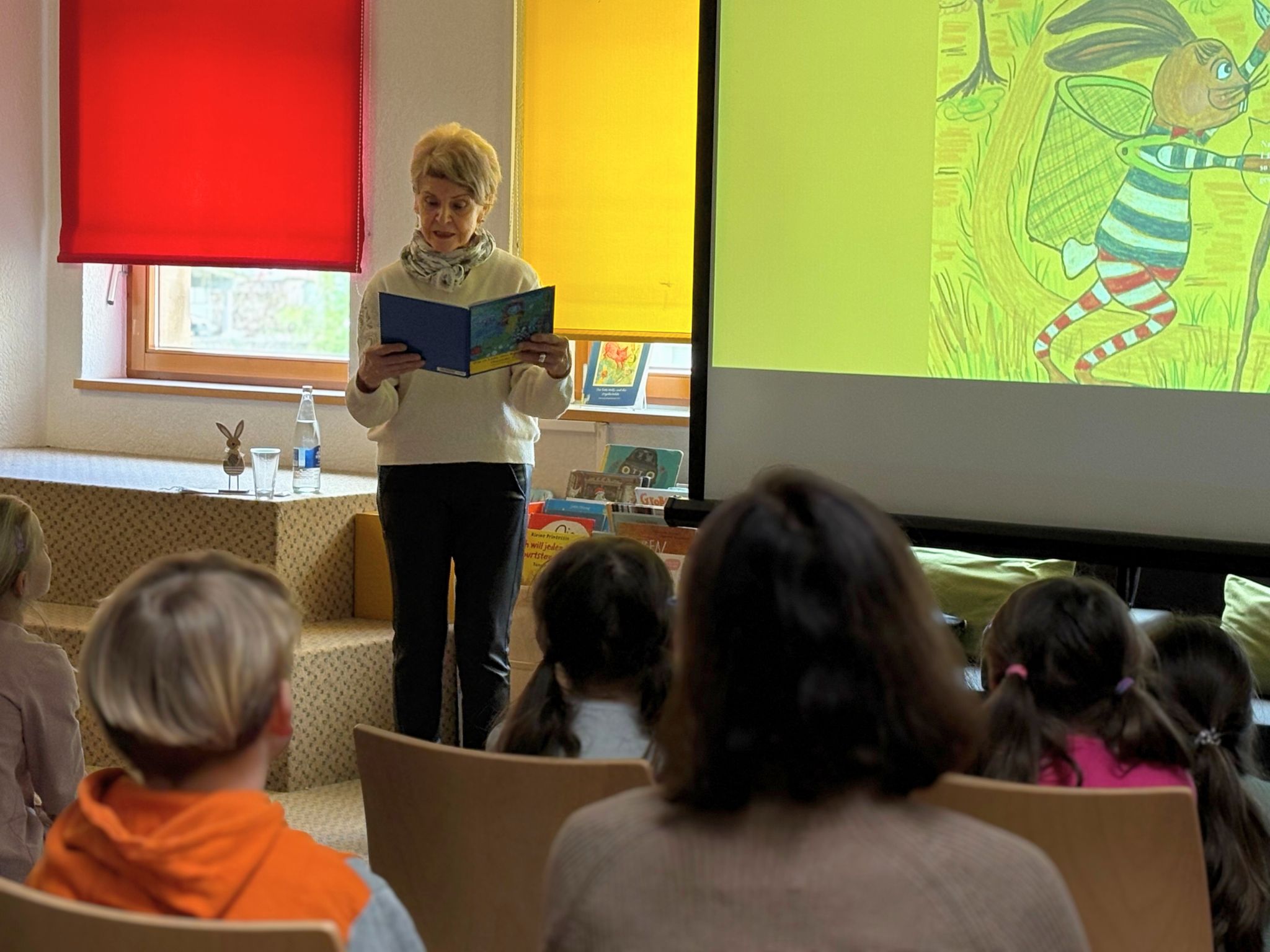 Eine Autorin liest aus einem Buch in der Mediathek vor. Viele Kinder sitzen davor und h&ouml;ren zu.
